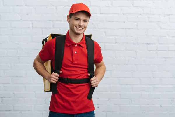 Delivery man in red uniform carrying thermo backpack and smiling at camera near brick wall — Stock Photo