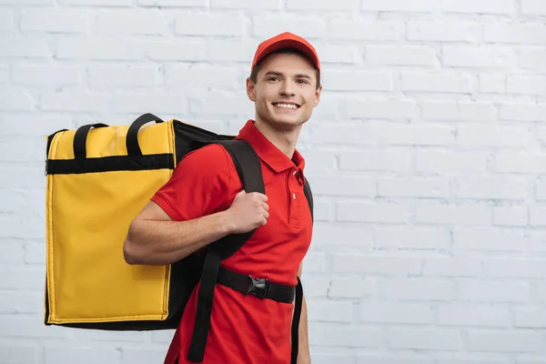 Livreur souriant avec sac à dos thermique regardant loin près du mur de briques — Photo de stock