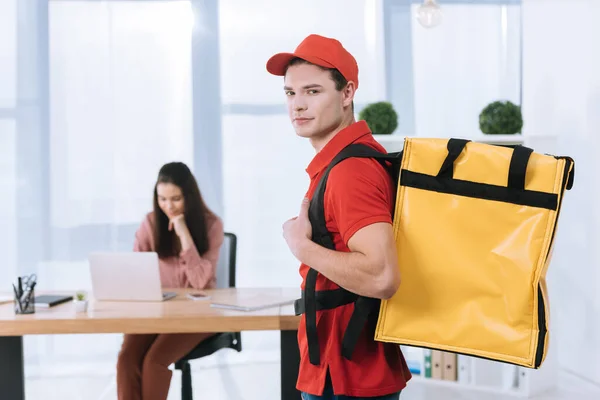 Selective focus of delivery man with thermo backpack looking at camera while businesswoman using laptop in office — Stock Photo