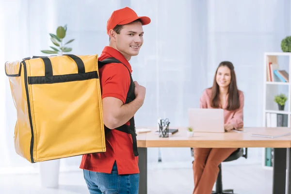 Selective focus of delivery man with thermo backpack smiling at camera with businesswoman at table in office — Stock Photo