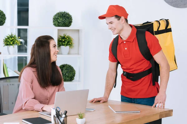 Empresaria sonriendo al mensajero con mochila térmica cerca de la mesa en la oficina - foto de stock