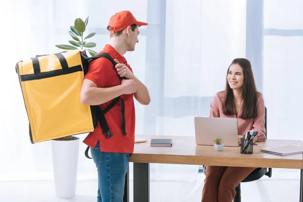 Attraktive Geschäftsfrau lächelt den Lieferanten mit Thermo-Rucksack an, während er am Tisch arbeitet — Stockfoto