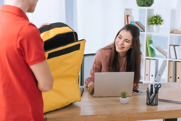 Selective focus of smiling businesswoman looking at courier with thermo bag at table in office — Stock Photo