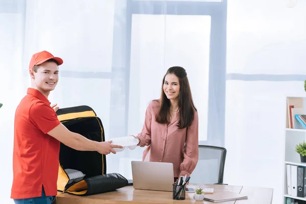 Delivery man with thermo bag giving food container to businesswoman and smiling at camera in office — Stock Photo