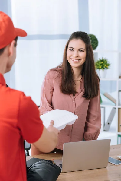 Foco seletivo de correio no uniforme vermelho que dá o container de comida à mulher de negócios sorridente à mesa — Fotografia de Stock