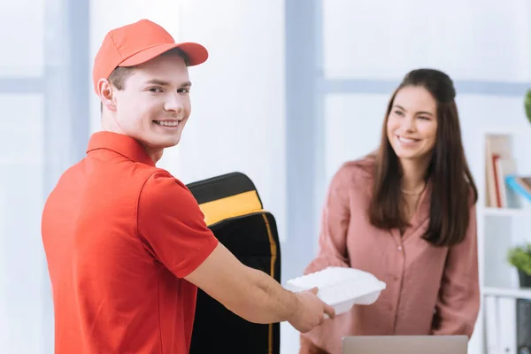 Selective focus of smiling courier looking at camera and giving takeaway box to smiling businesswoman in office — Stock Photo