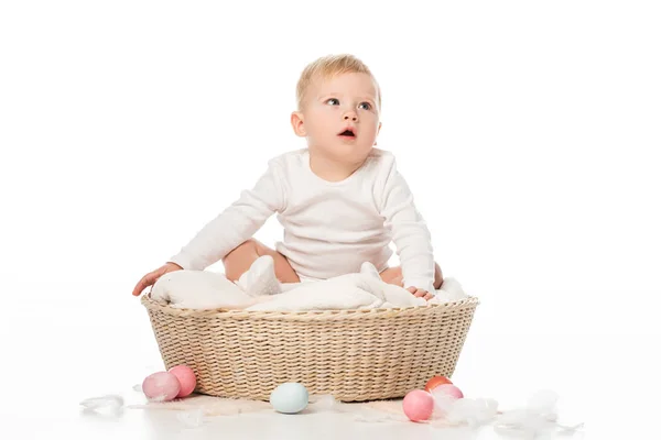 Criança com a boca aberta sentado em cobertor em cesta com ovos de Páscoa ao redor no fundo branco — Fotografia de Stock