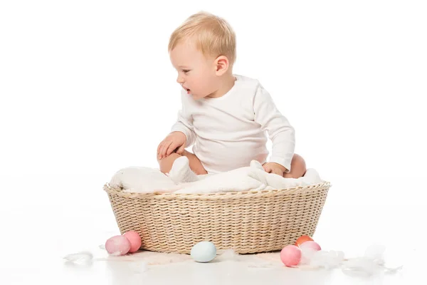 Niño mirando hacia abajo y sentado en la cesta con huevos de Pascua alrededor sobre fondo blanco - foto de stock