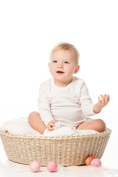 Niño con la mano levantada sentado en la cesta con huevos de Pascua alrededor sobre fondo blanco - foto de stock