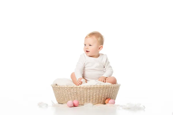 Kind mit offenem Mund schaut weg, sitzt im Korb neben Ostereiern auf weißem Hintergrund — Stockfoto