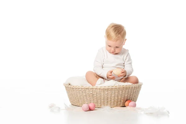 Niño con la cabeza baja sosteniendo huevo de Pascua, sentado en la manta en la cesta sobre fondo blanco - foto de stock