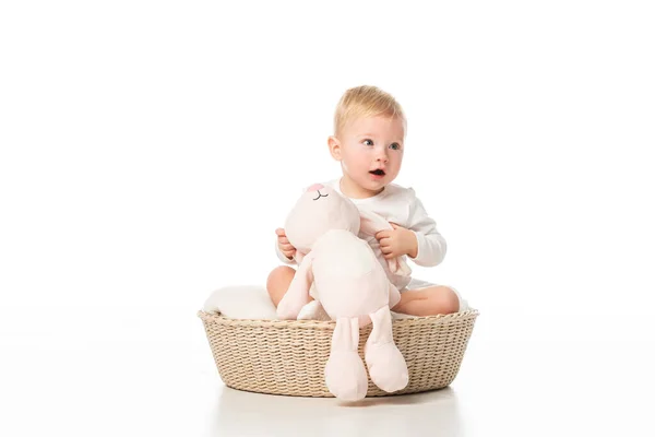 Милый ребенок с открытым ртом, держащий розового кролика и сидящий в корзине на белом фоне — стоковое фото