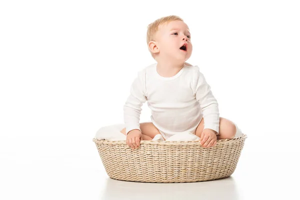 Niedlicher Junge, der mit offenem Mund wegschaut und im Korb auf weißem Hintergrund sitzt — Stockfoto