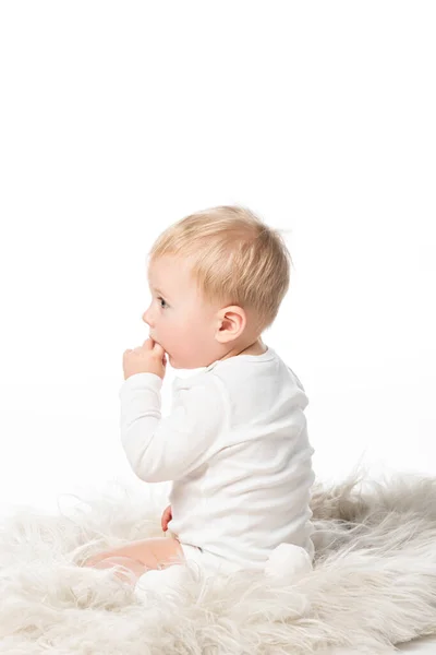 Vista laterale di bambino carino con dito in bocca, seduto su pelliccia isolata su bianco — Foto stock