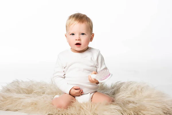 Criança olhando para a câmera com boca aberta segurando pente e sentado na pele isolada no branco — Fotografia de Stock