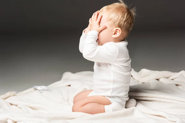 Side view of cute child covering face and kneeling on blanket isolated on black — Stock Photo