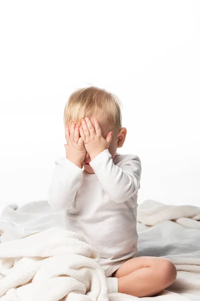 Bambino carino che copre il viso con le mani e seduto su una coperta isolata su bianco — Foto stock