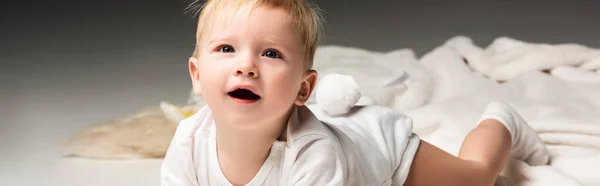 Niño acostado sobre manta, mirando hacia arriba con la boca abierta sobre fondo gris, plano panorámico - foto de stock