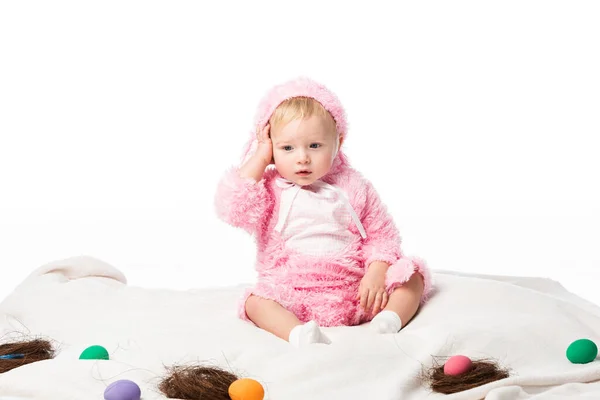 Upset child wearing rabbit costume, touching head on blanket with colorful decoration isolated on white — Stock Photo