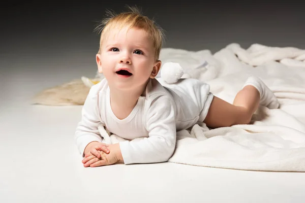 Enfant avec pompon sur culotte, couché sur la couverture, levant les yeux avec la bouche ouverte sur fond gris — Photo de stock