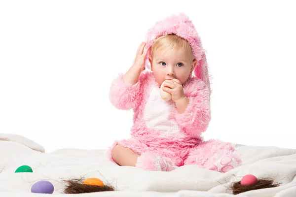 Niño con traje de conejo, poniendo huevo de Pascua en la boca, tocando la cabeza en la manta aislada en blanco - foto de stock