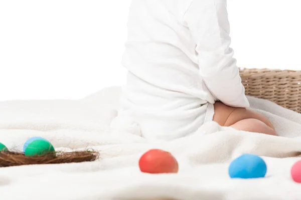 Vista cortada da criança sentada no cobertor ao lado da cesta e ninho com ovos de páscoa ao redor isolados em branco — Fotografia de Stock