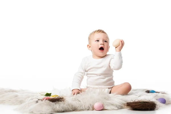Bambino a bocca aperta, con uovo di Pasqua in mano, seduto su pelliccia su sfondo bianco — Foto stock