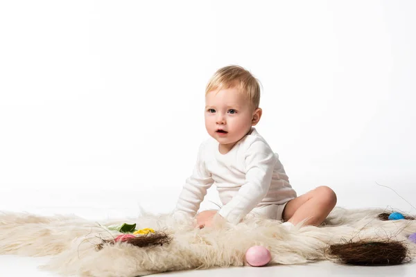 Child with open mouth, sitting on fur with nests and easter eggs around on white background — Stock Photo