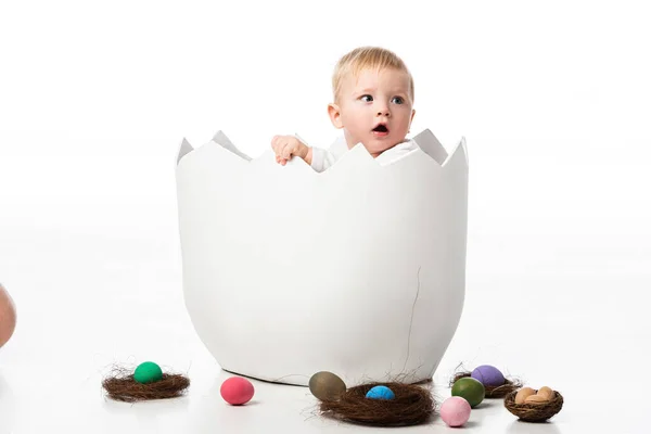Lindo niño con la boca abierta dentro de cáscara de huevo con nidos y huevos de Pascua sobre fondo blanco - foto de stock