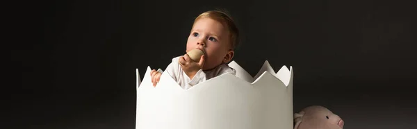 Lindo niño tomando huevo de Pascua a la boca por las manos apretadas dentro de la cáscara de huevo aislado en negro, tiro panorámico - foto de stock