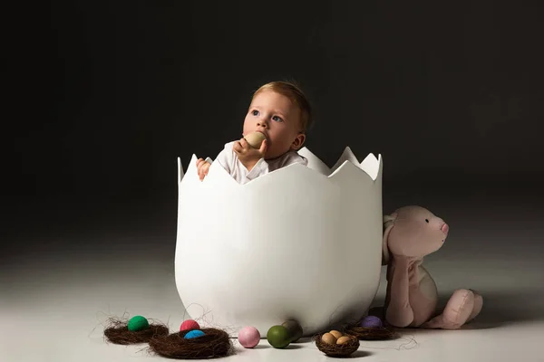 Niño tomando huevo de Pascua a la boca por las manos apretadas dentro de la cáscara de huevo sobre fondo negro - foto de stock