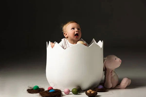 Child with open mouth looking up, holding Easter egg in eggshell on black background — Stock Photo