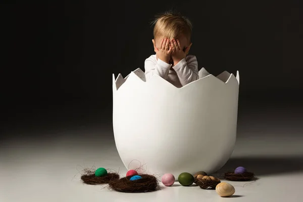 Enfant couvrant le visage à l'intérieur de la coquille d'oeuf à côté des œufs de Pâques et des nids sur fond noir — Photo de stock