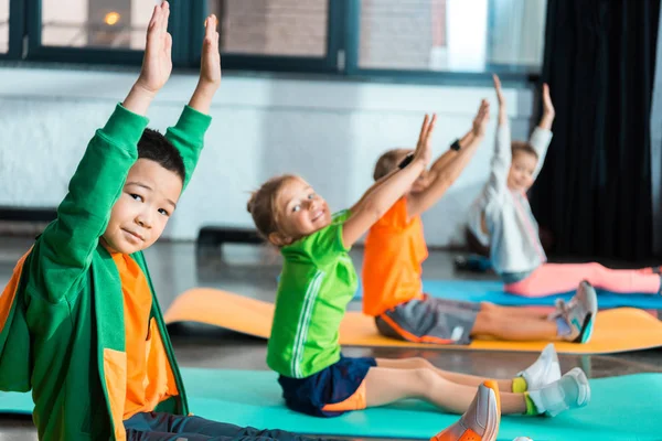 Enfoque selectivo de niños multiétnicos que miran a la cámara con las manos en el aire y hacen ejercicio en las alfombras de fitness en el gimnasio - foto de stock