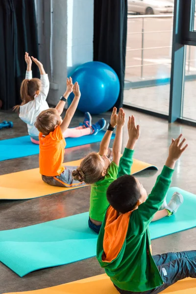 Vista de ángulo alto de niños multiétnicos con manos en el aire haciendo ejercicio en colchonetas de fitness en el gimnasio - foto de stock