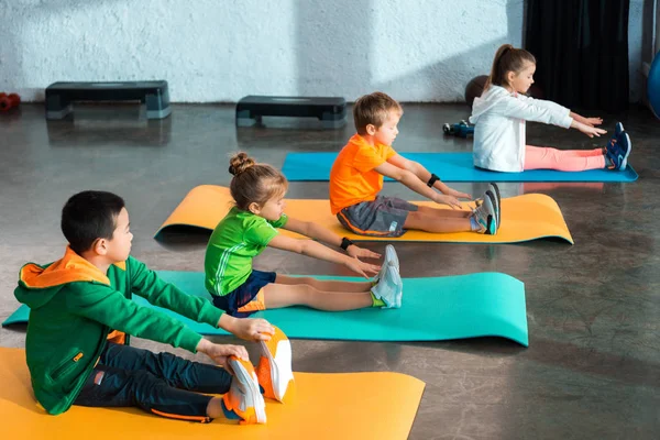 Bambini multiculturali che si allungano seduti su tappetini fitness in palestra — Foto stock