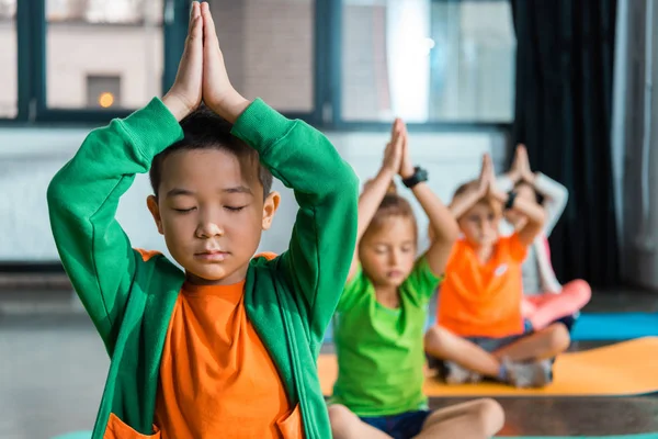 Selective focus of multicultural children meditating with clenched hands over heads in gym — Stock Photo