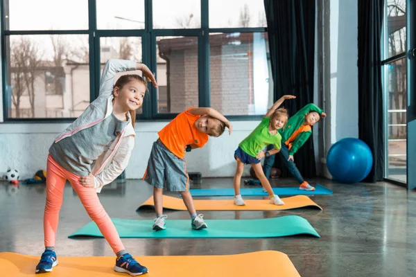 Enfoque selectivo de niños multiculturales calentándose en colchonetas de fitness en el centro deportivo - foto de stock
