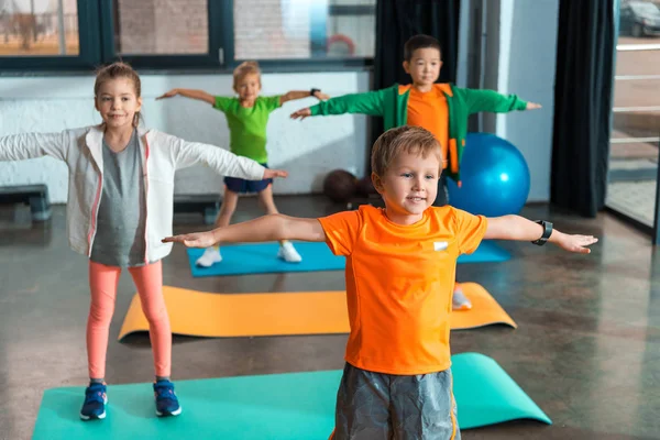 Selective focus of multiethnic children doing exercise with outstretched hands on fitness mats in gym — Stock Photo