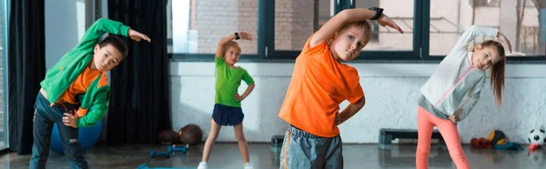 Selective focus of multicultural children warming up on fitness mats in gym, panoramic shot — Stock Photo