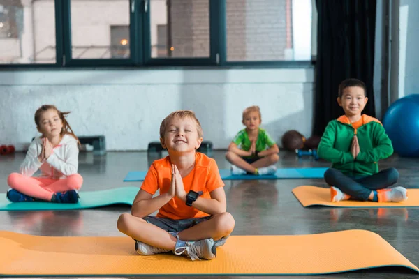 Селективный фокус мультиэтнических детей с сжатыми руками и скрещенными ногами, сидящих на фитнес-ковриках — стоковое фото