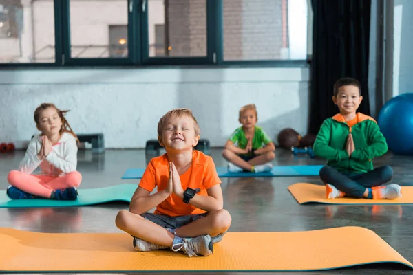 Enfoque selectivo de niños multiétnicos con las manos apretadas y las piernas cruzadas sentados en colchonetas de fitness — Stock Photo
