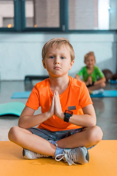 Concentration sélective du garçon avec les mains serrées et les jambes croisées sur le tapis de fitness dans le centre sportif — Photo de stock