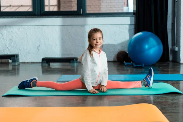 Foco seletivo da criança sorrindo e fazendo split no tapete de fitness no ginásio — Fotografia de Stock