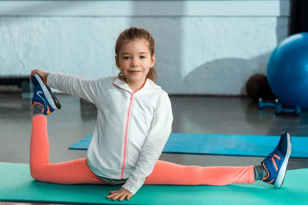 Enfoque selectivo del niño sonriendo, estirándose, haciendo split en la estera de fitness en el gimnasio - foto de stock