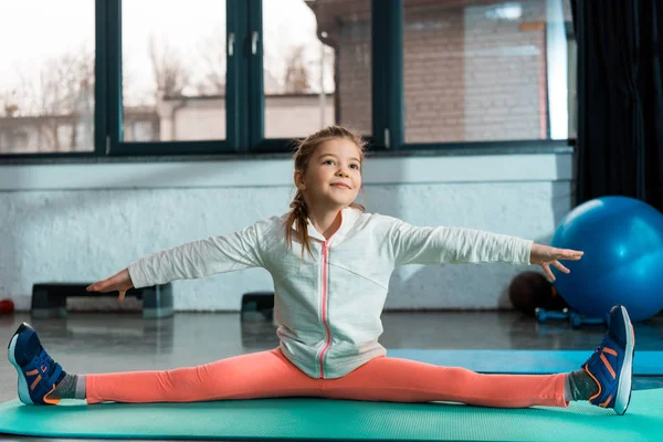 Selective focus of positive child with outstretched hands doing split on fitness mat in gym — Stock Photo