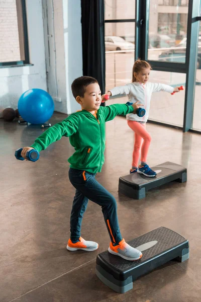 Vista de alto ângulo de crianças multiétnicas com as mãos estendidas segurando halteres e fazendo Step Aerobics no ginásio — Fotografia de Stock