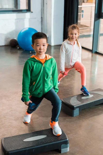 Hohe Blickwinkel auf multiethnische Kinder, die Hanteln in der Hand halten und auf Trittbrettern in der Turnhalle trainieren — Stockfoto