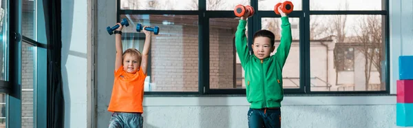 Front view of multicultural kids raising hands with dumbbells while doing exercise in gym, panoramic shot — Stock Photo