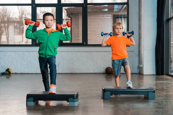 Vue de face d'enfants multiculturels tenant des haltères et faisant de l'aérobic pas à pas dans une salle de gym — Photo de stock